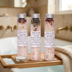 Three Soma & Sage botanical bath salt tubes with an unlit tealight candle and a folded ivory towel on an oak tray spanning a bathtub.