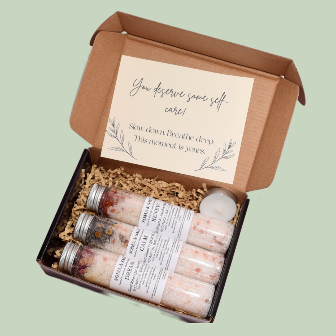 Open kraft gift box containing Dream, Calm, Renew bath salt tubes, shredded paper filler, an unlit tealight candle, a printed self-care message.