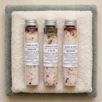 Three Soma & Sage bath salt tubes labelled Dream, Calm, Renew, lying side by side on plush ivory and sage-green towels.