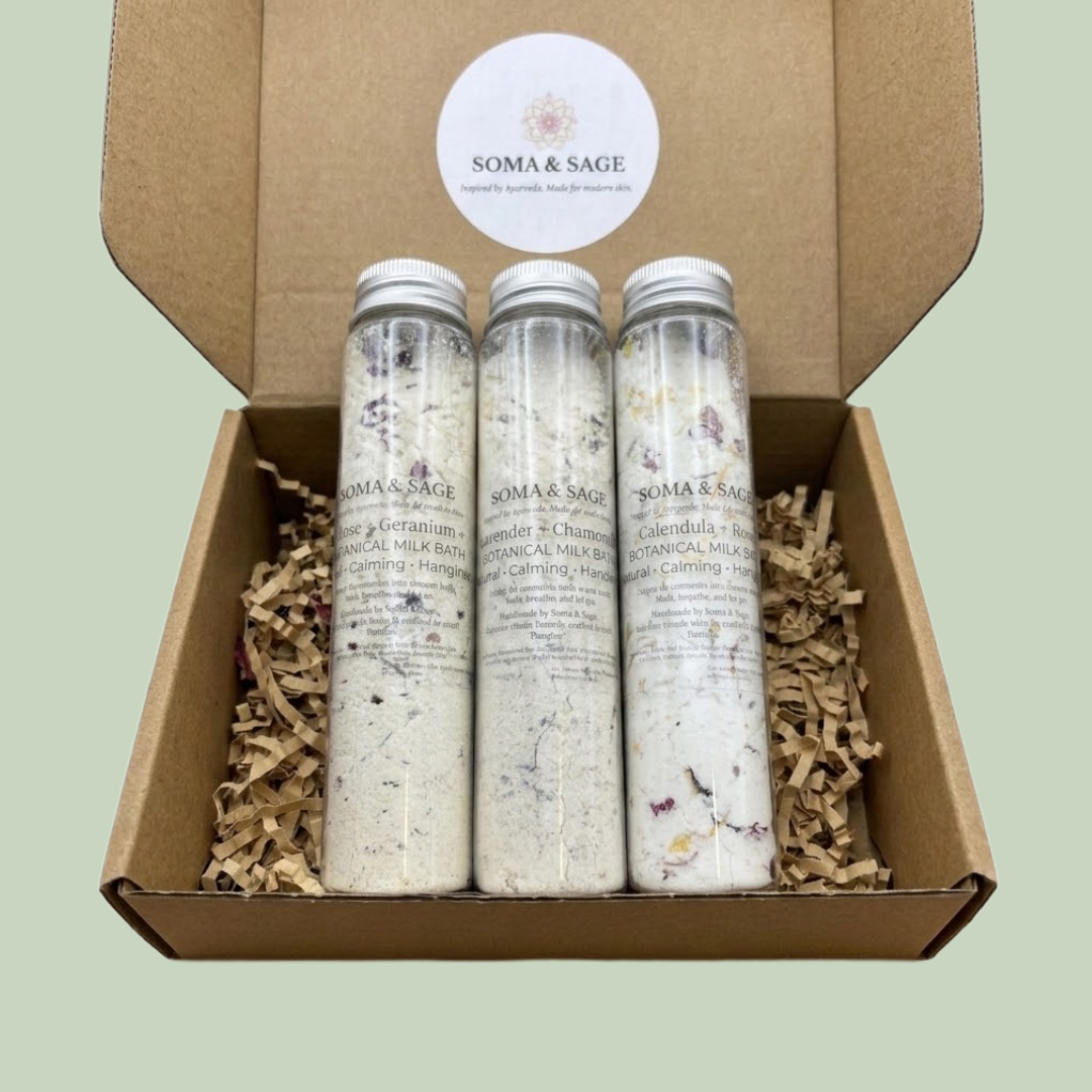 Three Soma & Sage botanical milk bath bottles with silver caps nestled in shredded paper inside an open kraft gift box.