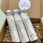 Three Soma & Sage botanical milk bath bottles with silver caps nestled in shredded paper inside an open kraft gift box.