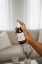 Hand holding Lavender Unwind amber glass room spray in a bright, airy living room with neutral linen sofa and sheer curtains — Sensory for Calm